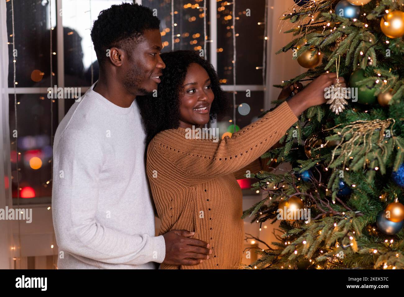 Lächelnder junger afroamerikanischer Kerl und Frau umarmt und schmückt Weihnachtsbaum mit Spielzeug Stockfoto
