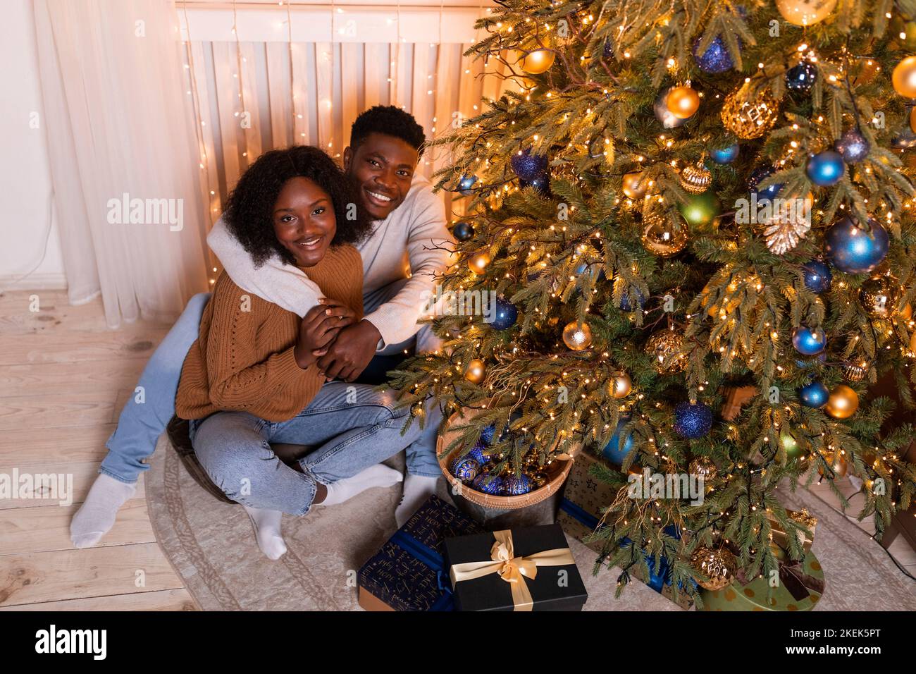 Lächelndes, tausendjähriges schwarzes Paar umarmt sich und sitzt auf dem Boden, schmückt Weihnachtsbaum mit Lichtern, Spielzeug Stockfoto