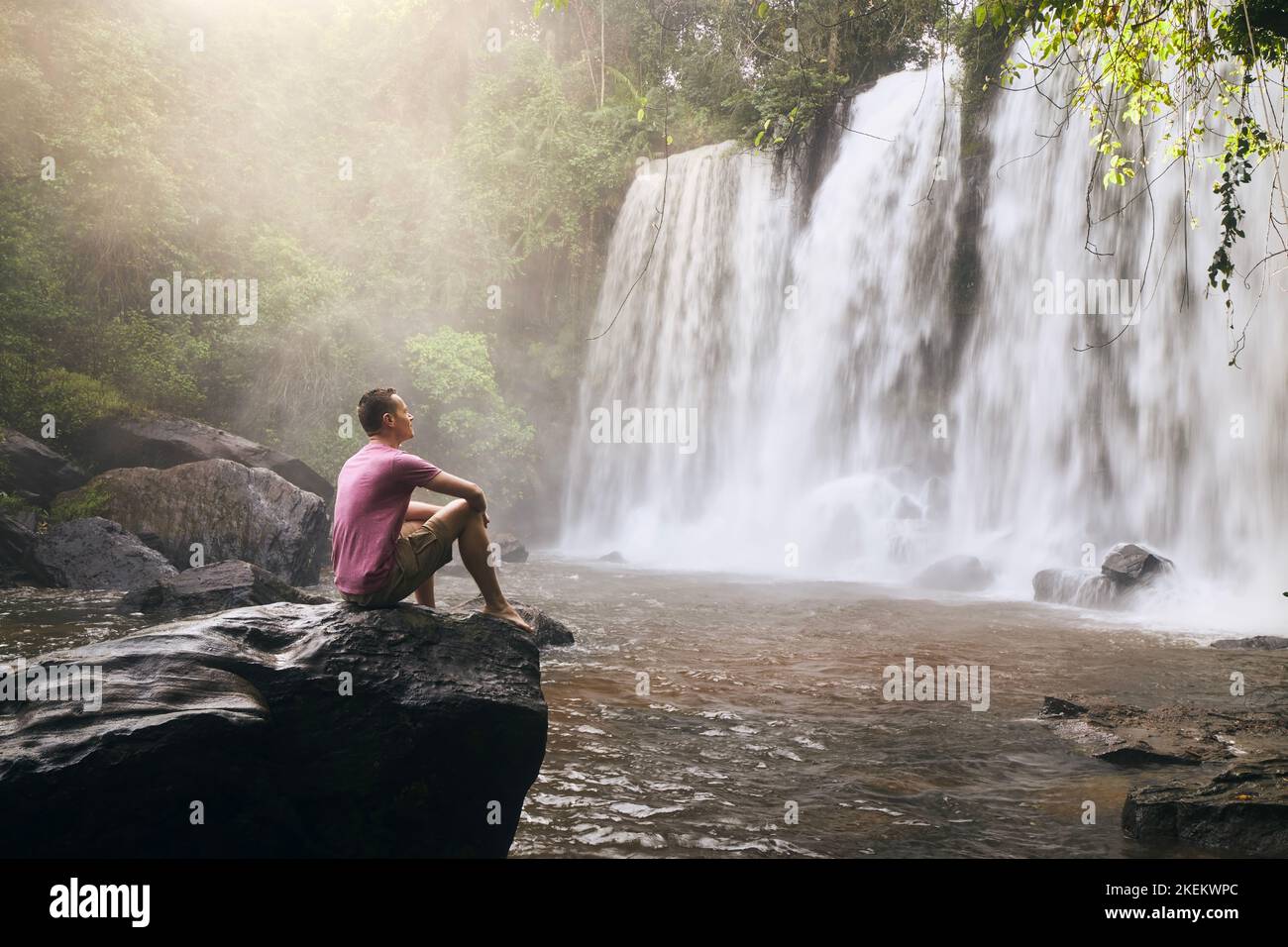 Mann sitzt auf einem Felsen vor einem hohen Wasserfall in den Bergen in der tropischen Landschaft von Camobodia. Stockfoto