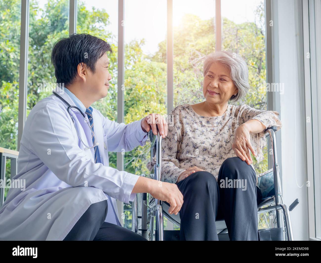 Lächelnd positiver asiatischer Mann Arzt im weißen Mantel spricht mit Sorgfalt zu älteren älteren älteren weiblichen Patienten, die in einem Rollstuhl in der medizinischen Praxis in der Zusammenarbeit sitzen Stockfoto