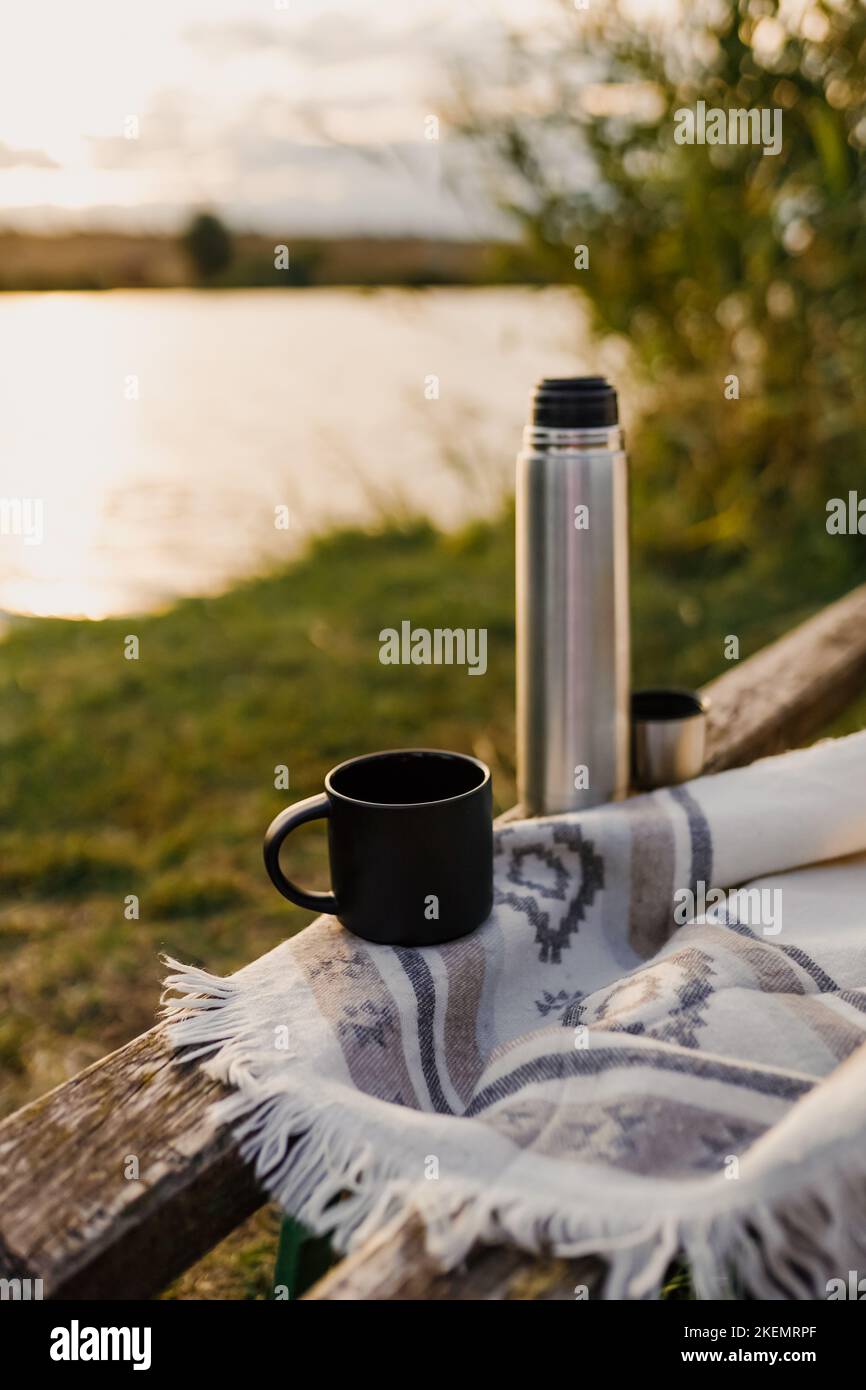 Toller Sonnenuntergang am See, Kaffee, Romantik. Ästhetisches Picknick im Herbst. Eine schöne Decke auf einer Bank vor dem See. Ästhetisches Picknick im Freien. Stockfoto