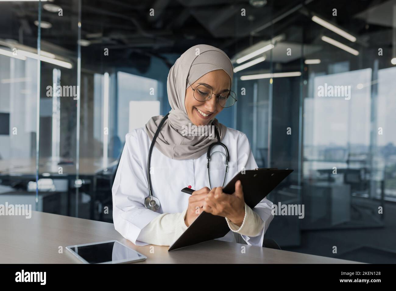 Eine junge schöne arabische Ärztin, eine muslimische Frau, sitzt in einem Hijab mit einem Stethoskop im Büro am Tisch. Arbeitet mit Dokumenten, zeichnet die Krankengeschichte von Patienten auf. Stockfoto