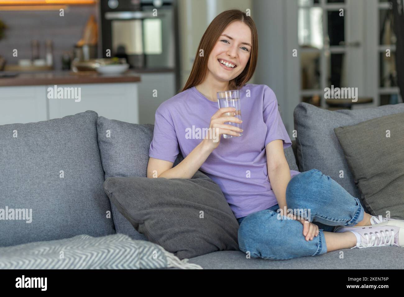 Schöne junge kaukasische Frau hält ein Glas frisches sauberes Wasser auf der Couch zu Hause sitzen, lächelt freundlich Stockfoto