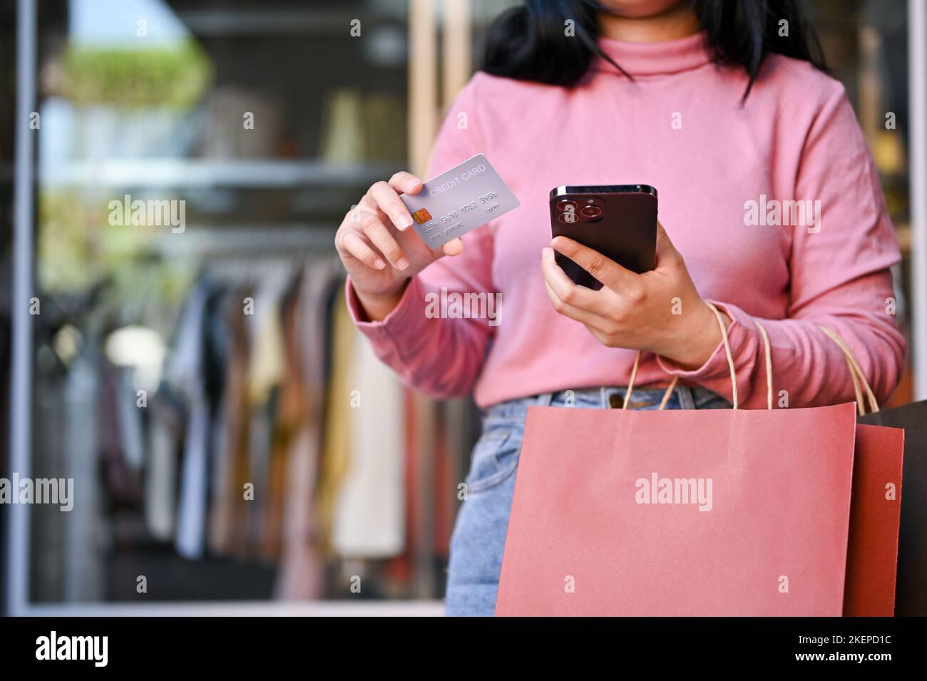 Die junge Asiatin mit ihren Einkaufstaschen, die eine Kreditkarte und ihr Smartphone in der Hand halten, steht vor einem Kleiderladen. Stockfoto