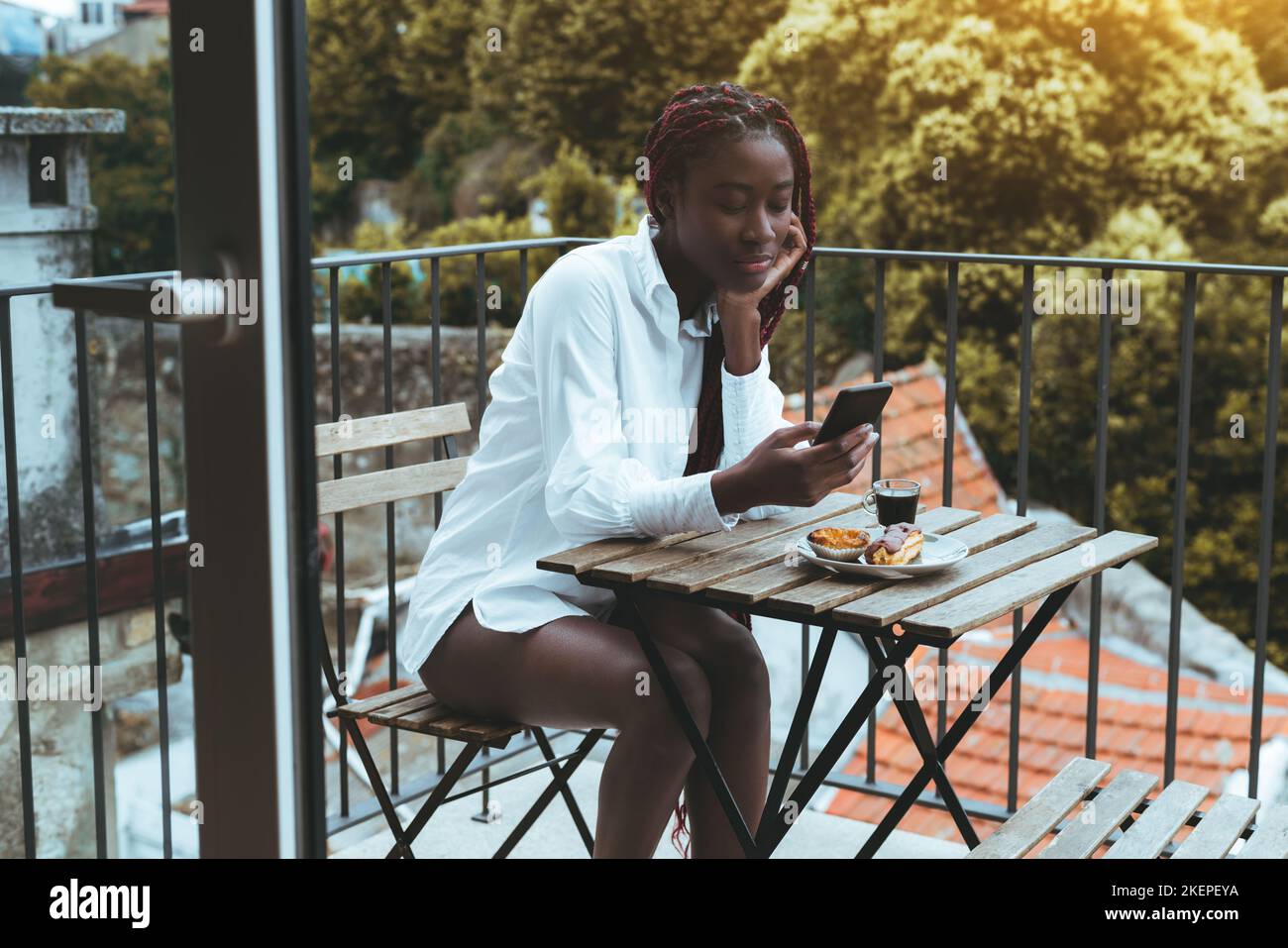 Eine schlaffe junge schwarze Frau mit roten Zöpfen scrollt faul einen Social-Media-Feed auf ihrem Smartphone, während sie mit einem Espresso-Kaffee frühstückt Stockfoto