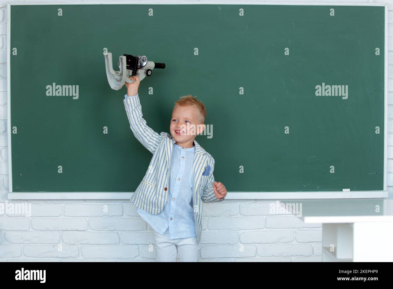 Kind in der Schule. Das Kind lernt im Unterricht vor dem Hintergrund der Tafel. Frühzeitige Bildung. Kleines Kind mit Mikroskop. Stockfoto