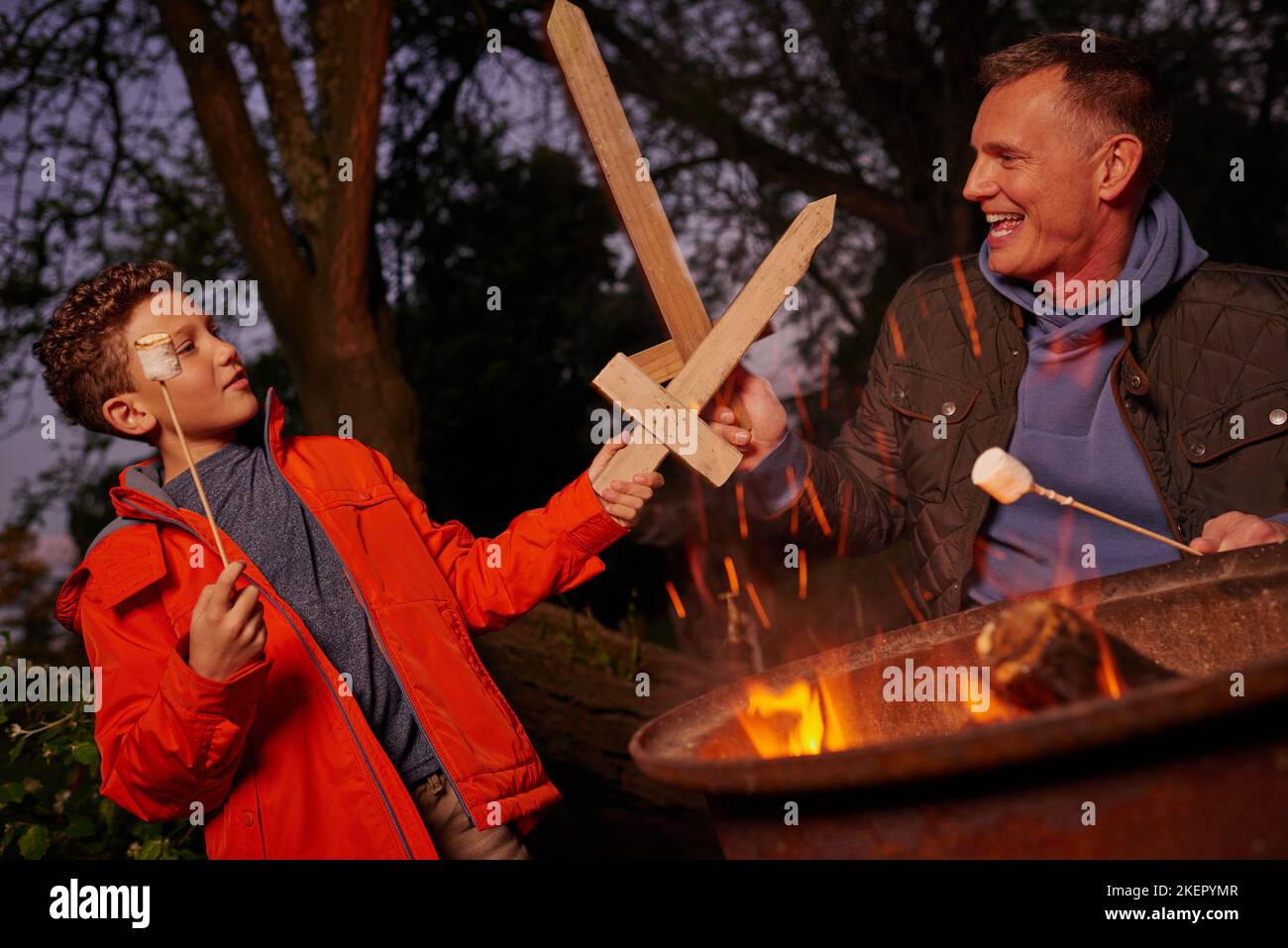 Ein Vater und ein Sohn spielen mit Holzschwertern, während sie mit Marschwalben über einem Feuer toasten. Stockfoto