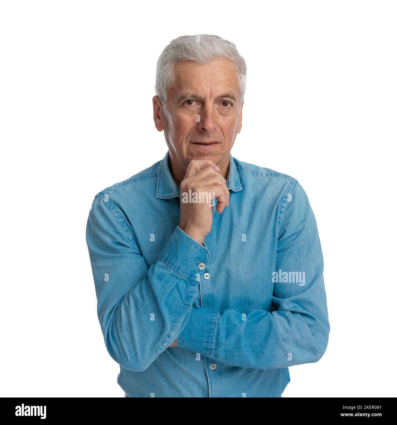 Nachdenklicher alter Mann im Jeanshemd, der die Arme zusammenklappte, das Kinn berührte, vor weißem Hintergrund im Studio dachte und posierte Stockfoto