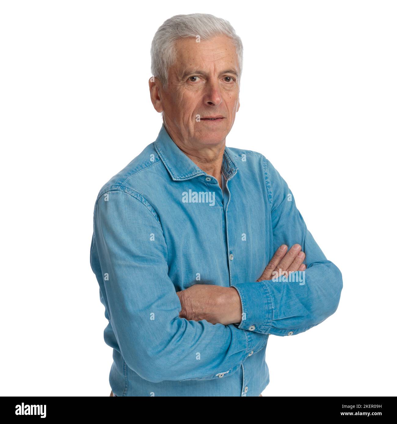 Portraitbild des selbstbewussten alten Mannes in Denim-Hemd, das Arme zusammenklappte und vor weißem Hintergrund im Studio posierte Stockfoto