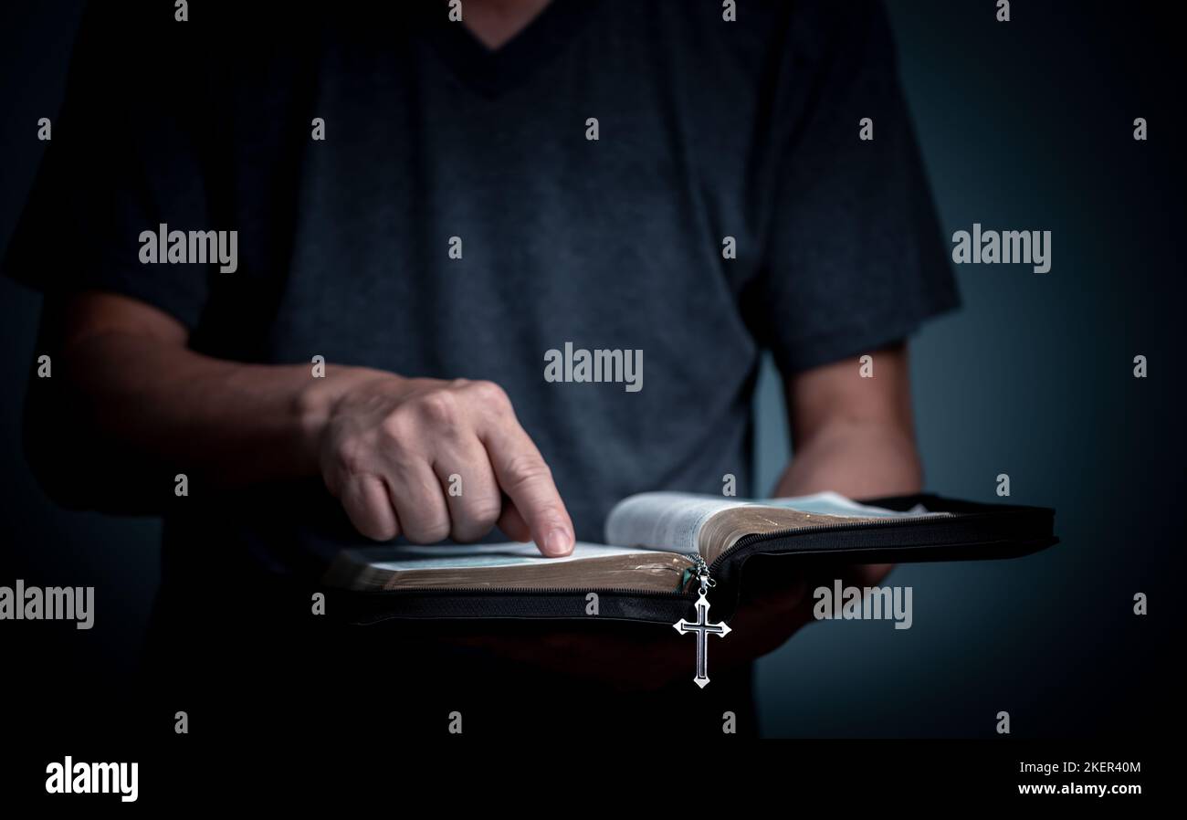 Junger Mann, der die Heilige Bibel liest und auf das Wort mit einer Kreuzkette im Schriftkegel des Buches zeigt. Stockfoto