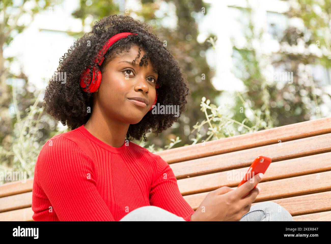 Junge Frau, die auf einer Bank sitzt und Musik mit Kopfhörern hört, mit einem Ausdruck von Selbstbewusstsein im Freien Stockfoto