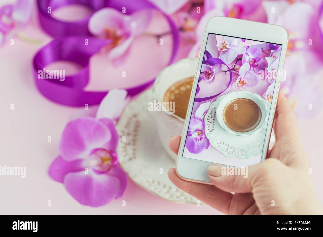 Frau, die mit ihrem Smartphone ein schönes Bild von einer Tasse Kaffee mit rosa Orchideen und violetten Bändern auf dem Tisch gemacht hat. Stockfoto