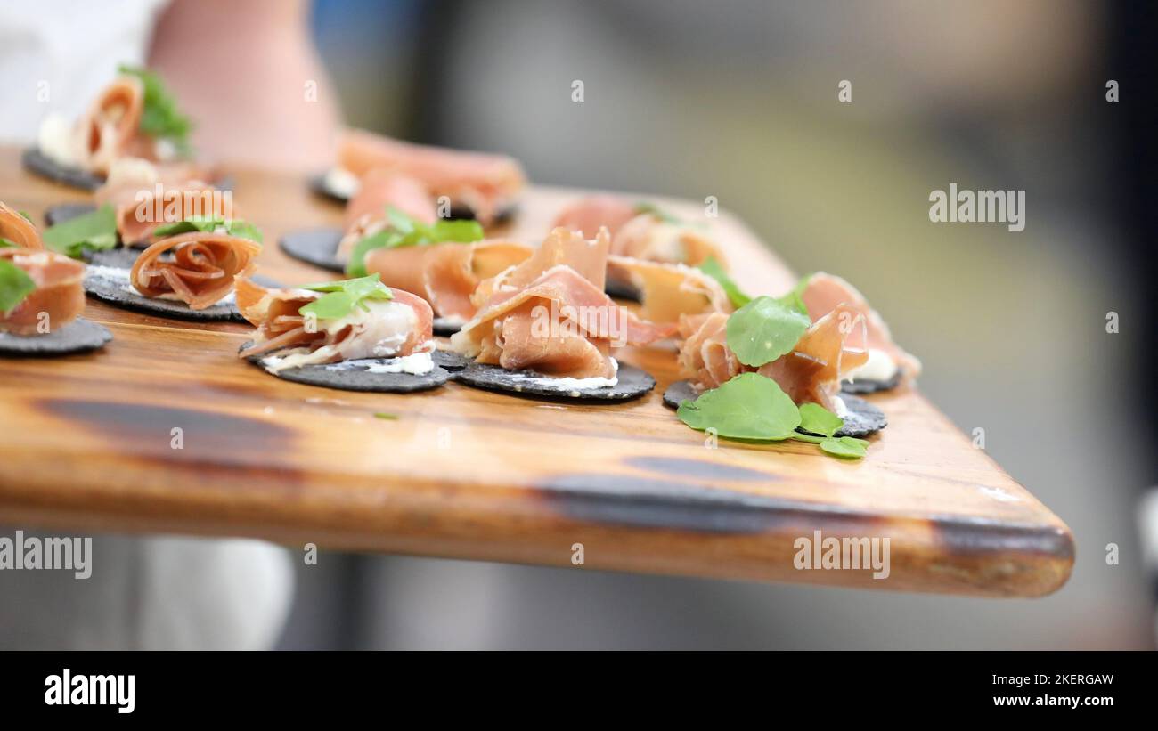 Eine rustikale Holzplatte mit Häppchen oder Fingerfood. Schwarze Wafer Cracker mit Käse und geräuchertem Fleisch wie Prosciutto gerollt und kleinen grünen Blatt Stockfoto