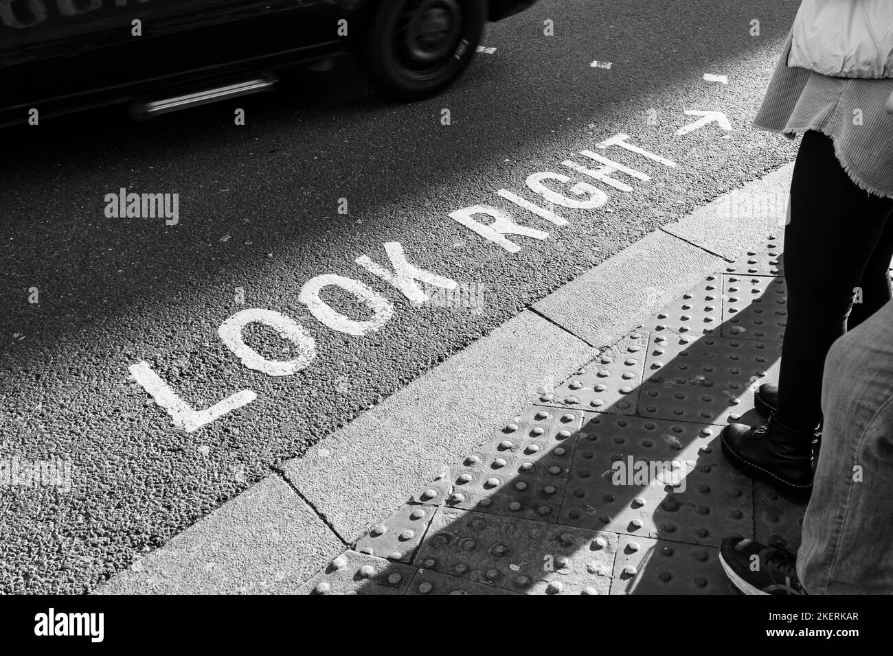 Ikonische Straßenmarkierung „Look Right“ für Fußgänger in London mit einem vorbeifahrenden Auto. Konzept: Überqueren der Straße, Verkehrssicherheit, Passanten warten Stockfoto