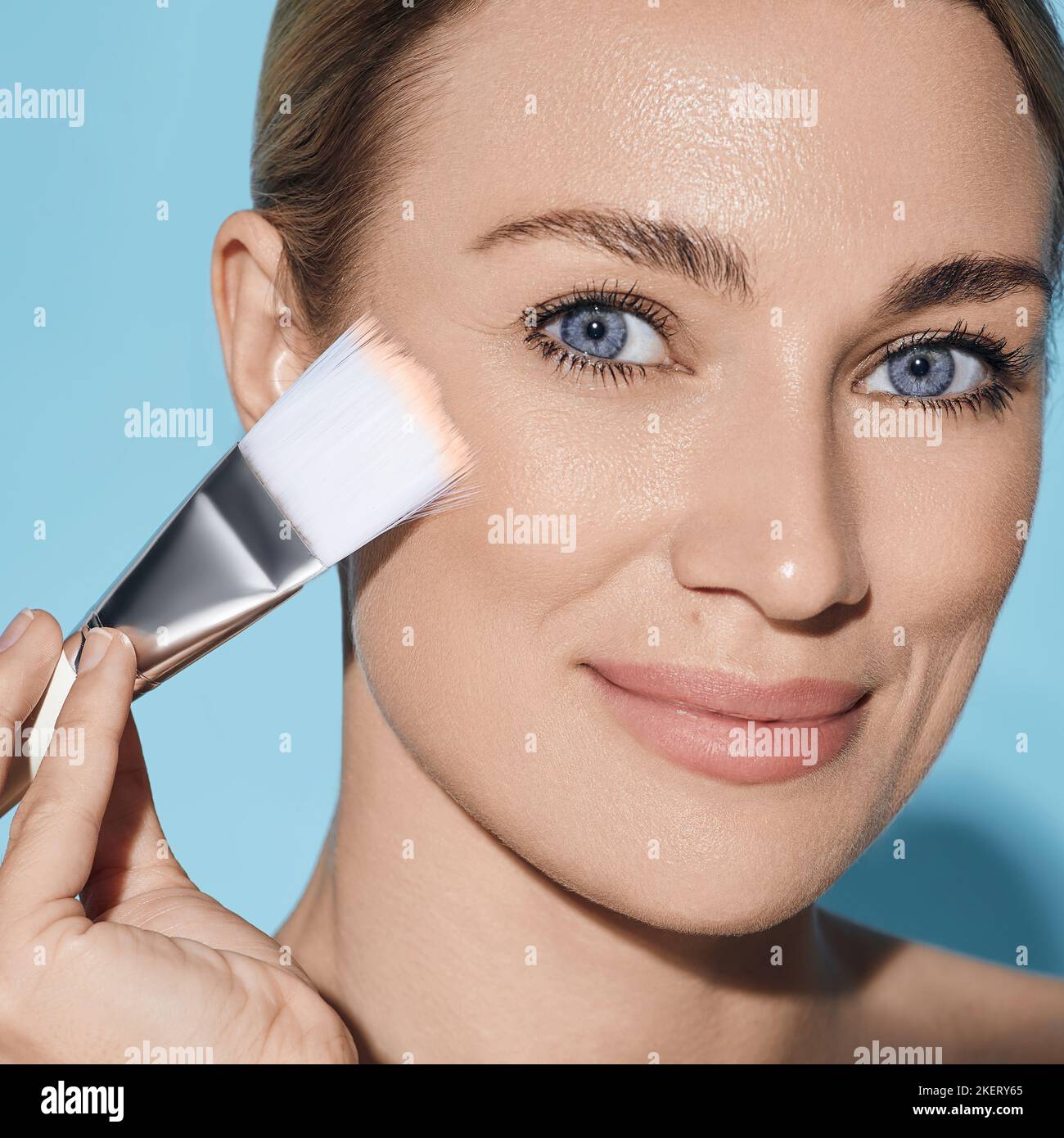 Schöne Frau, die Toncreme auf ihr perfektes Gesicht mit kosmetischem Pinsel aufträgt, isoliert auf blauem Hintergrund. Gesichtskosmetik, Foundation Stockfoto