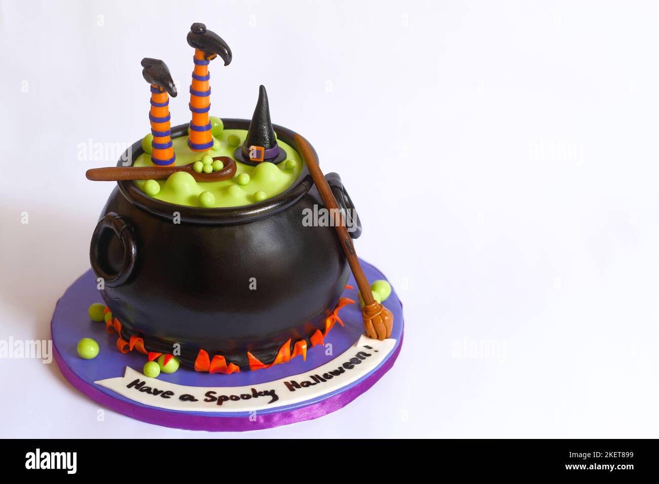 Kreativer und gruseliger Halloween-Hexenkessel-Kuchen auf weißem Hintergrund. Hexenhut, Beine und ein Holzlöffel auf einem schwarzen Topf mit kochender grüner Flüssigkeit. Stockfoto