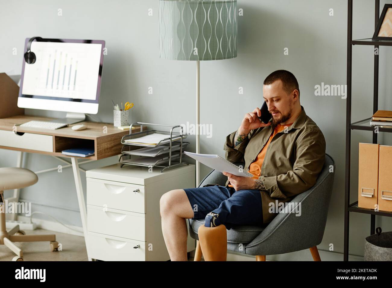 Porträt eines Mannes mit prothetischen Beinanrufen per Telefon, während er auf einem Stuhl im Home-Office-Innenraum sitzt, kopieren Raum Stockfoto