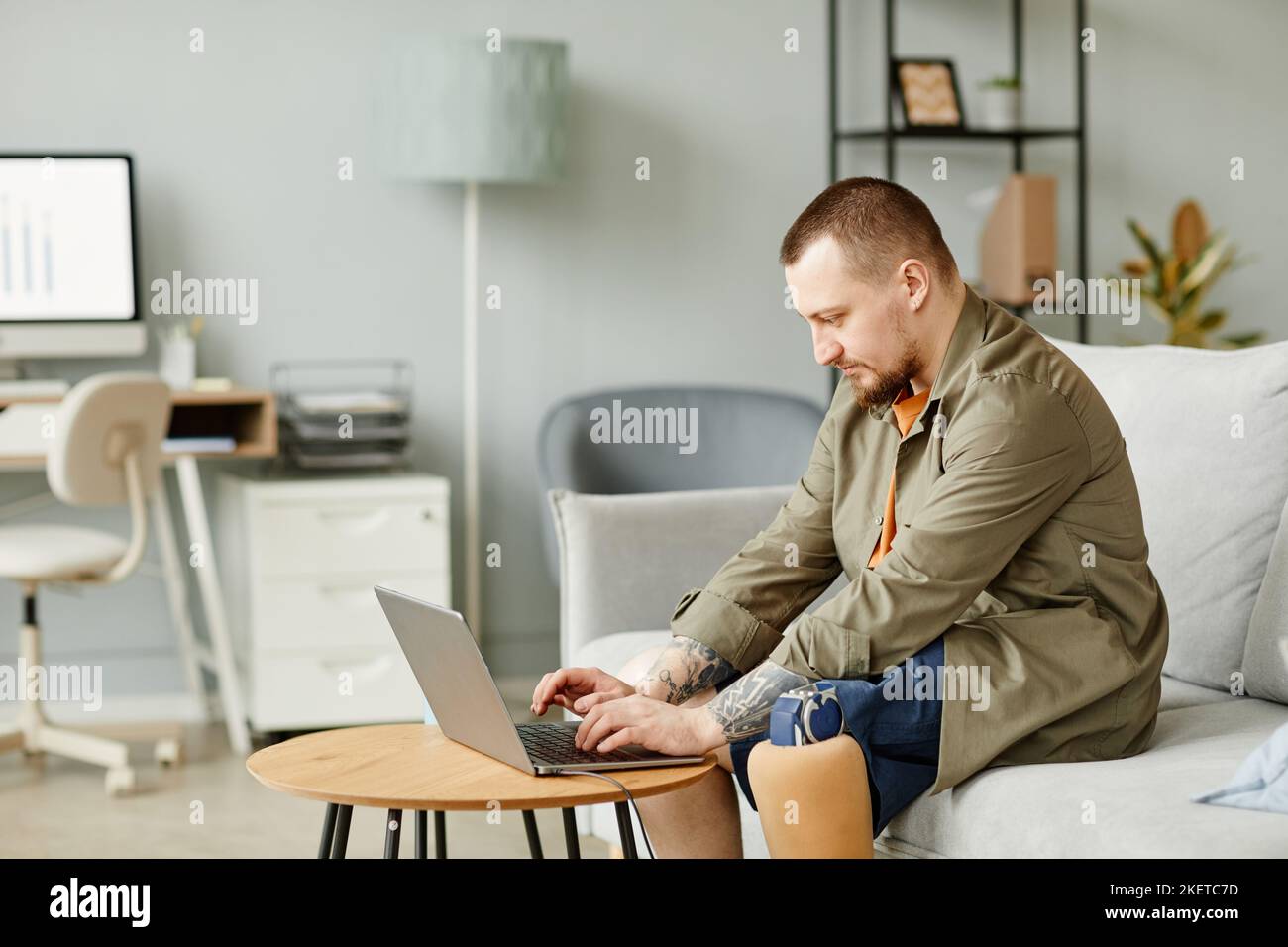 Seitenansicht Porträt eines Mannes mit Beinprothese mit Laptop, während er in minimalem Büroinnenraum arbeitet, Kopieplatz Stockfoto