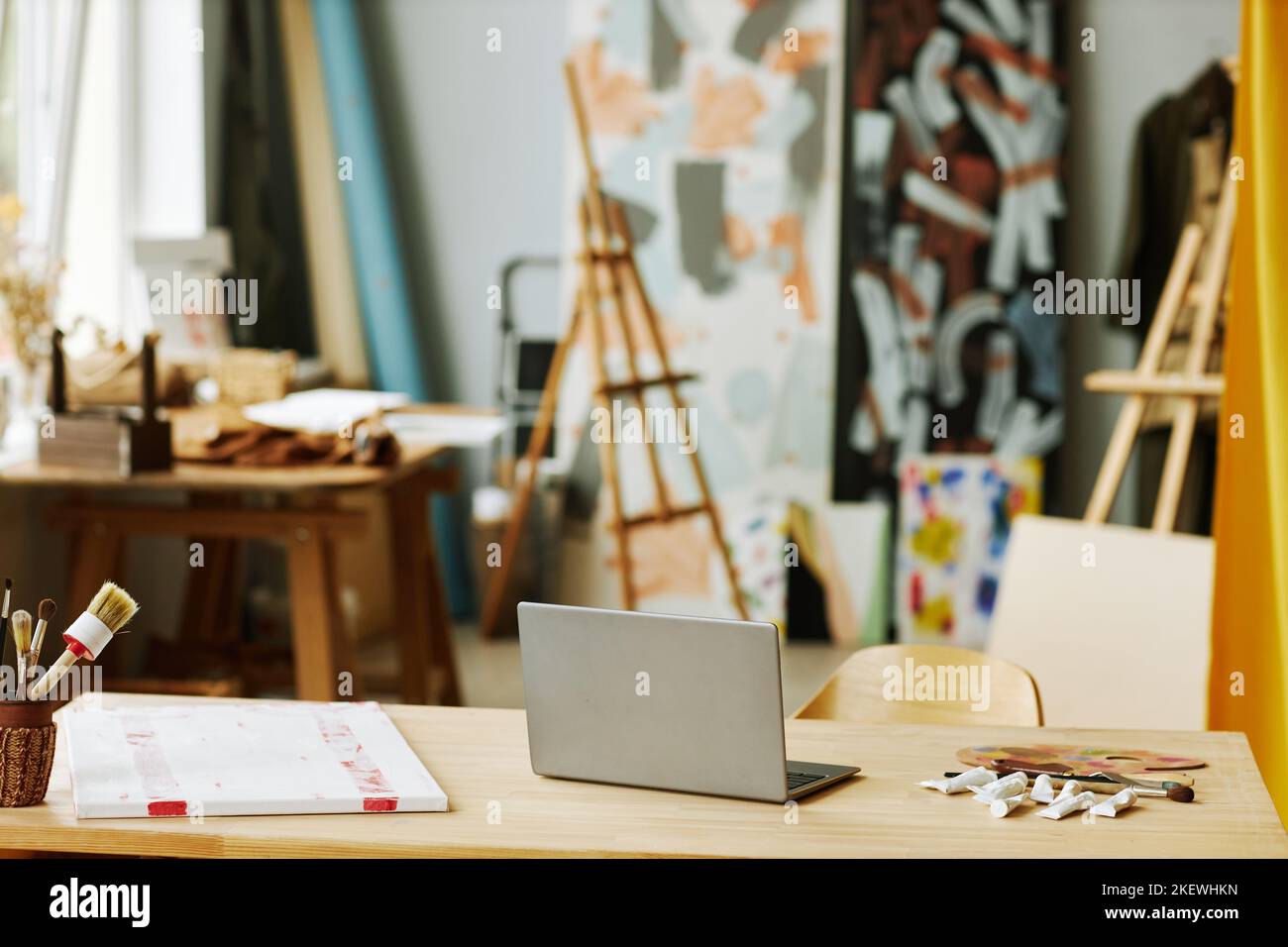 Laptop, der zwischen vielen Pinsel, Farbpalette, weißer Leinwand und mehreren Tuben mit Acrylfarben auf dem Schreibtisch steht Stockfoto