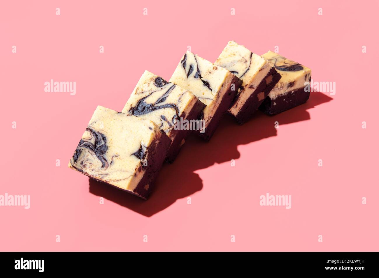 Hausgemachter, geschichteter Kuchen mit einer Schicht Schokolade und einem Käsekuchen. Brownie-Scheiben mit Käsekuchen in hellem Licht auf einem rosafarbenen Tisch Stockfoto