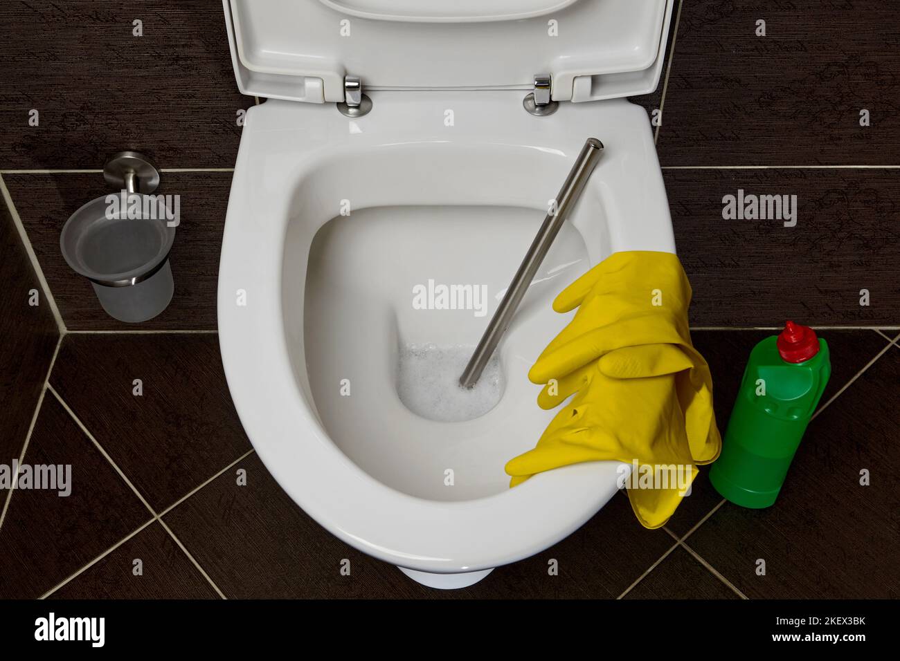 Toilettenschüssel mit Toilettenbürste, gelben Gummihandschuhen und einem Reinigungs- und Desinfektionsmittel auf dem Hintergrund dunkler Keramikfliesen im Toilettenraum Stockfoto