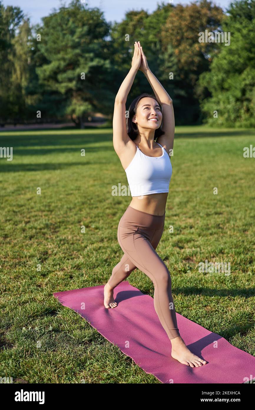 Vertikale Aufnahme eines jungen asiatischen Fitness-Mädchens, das Yoga Asana macht, in Baumhaltung steht und lächelt und im Park auf einer Gummimatte ausübt Stockfoto