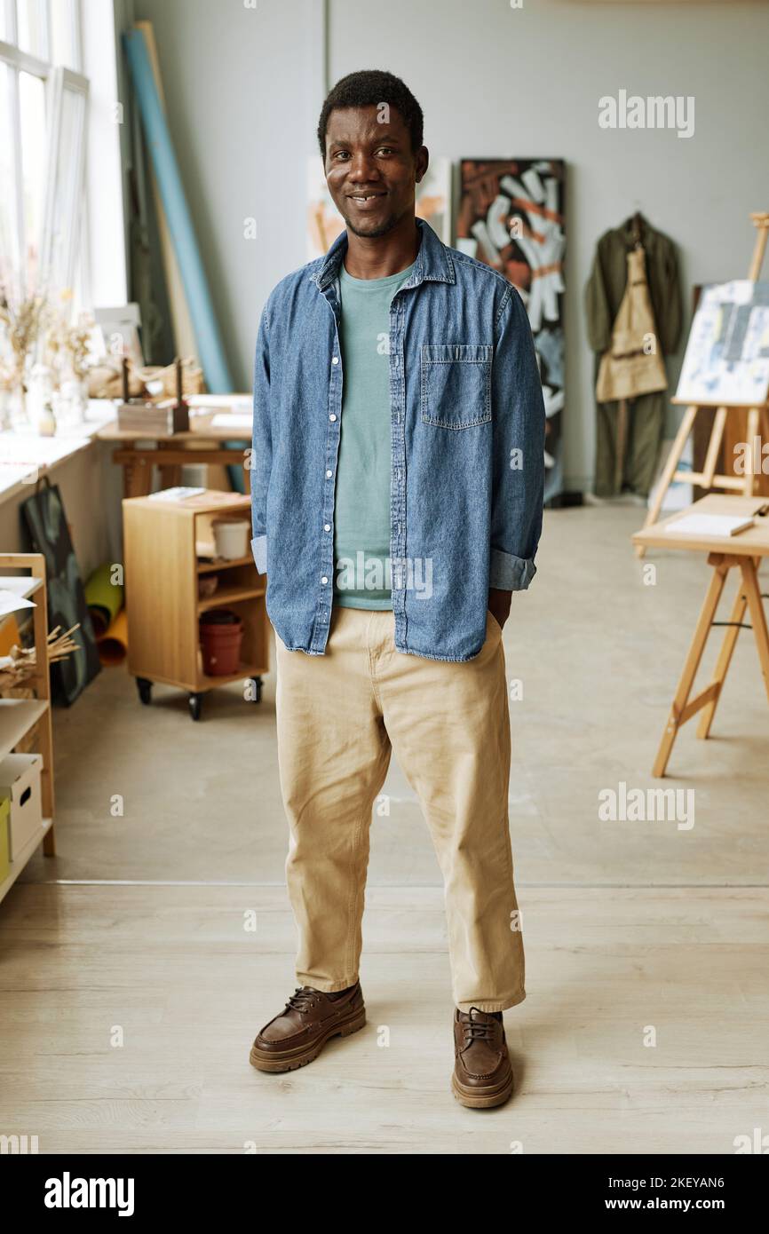 Junger lächelnder afroamerikanischer Mann in Casualwear, der vor der Kamera im geräumigen Kunststudio oder in der Schule der Malerei steht Stockfoto