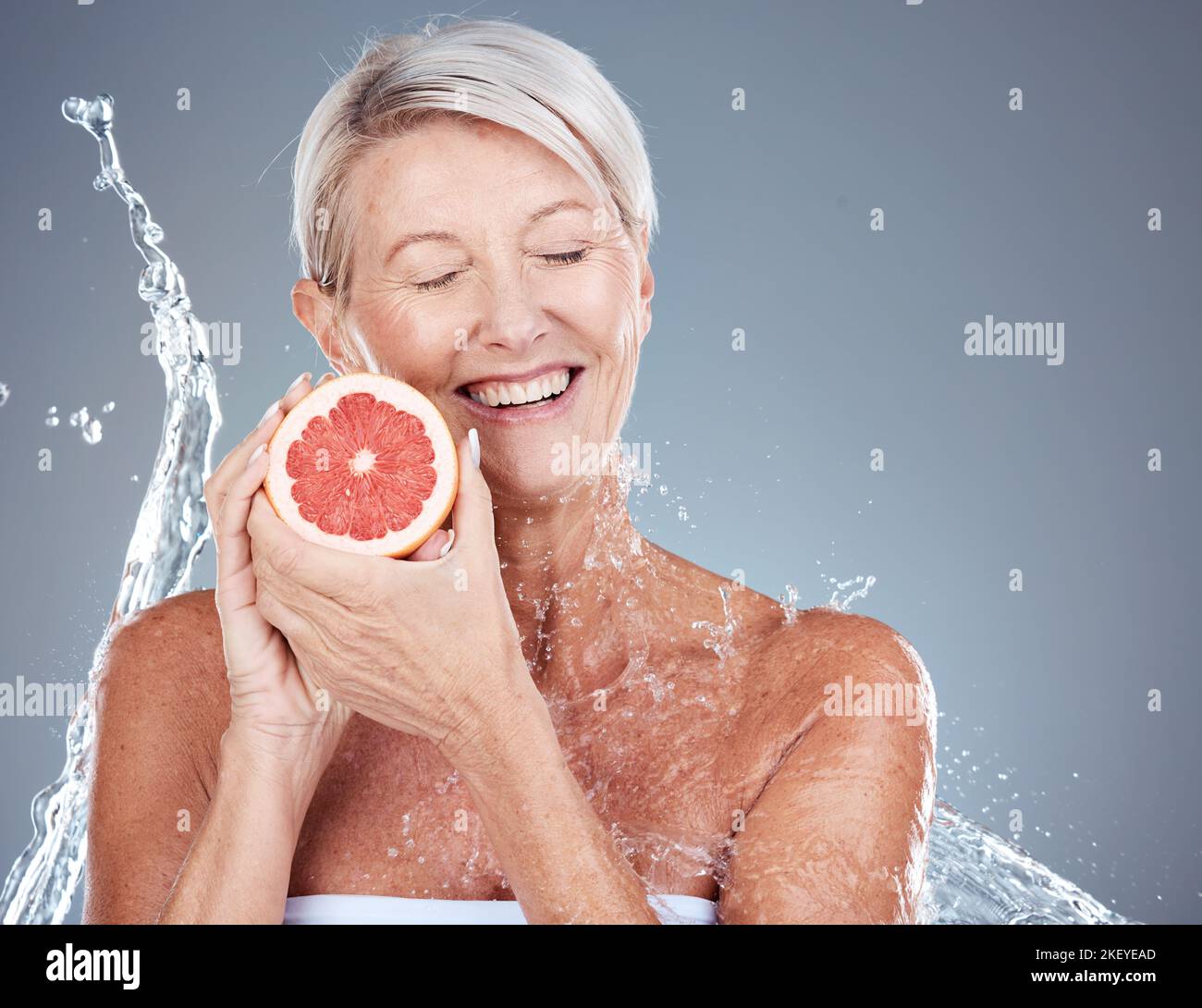 Hautpflege, Wasser und Porträt einer alten Frau mit Grapefruit in grauem Hintergrund Studio für Wellness. Schönheit, Wasserspritzer und ältere Weibchen mit Früchten Stockfoto