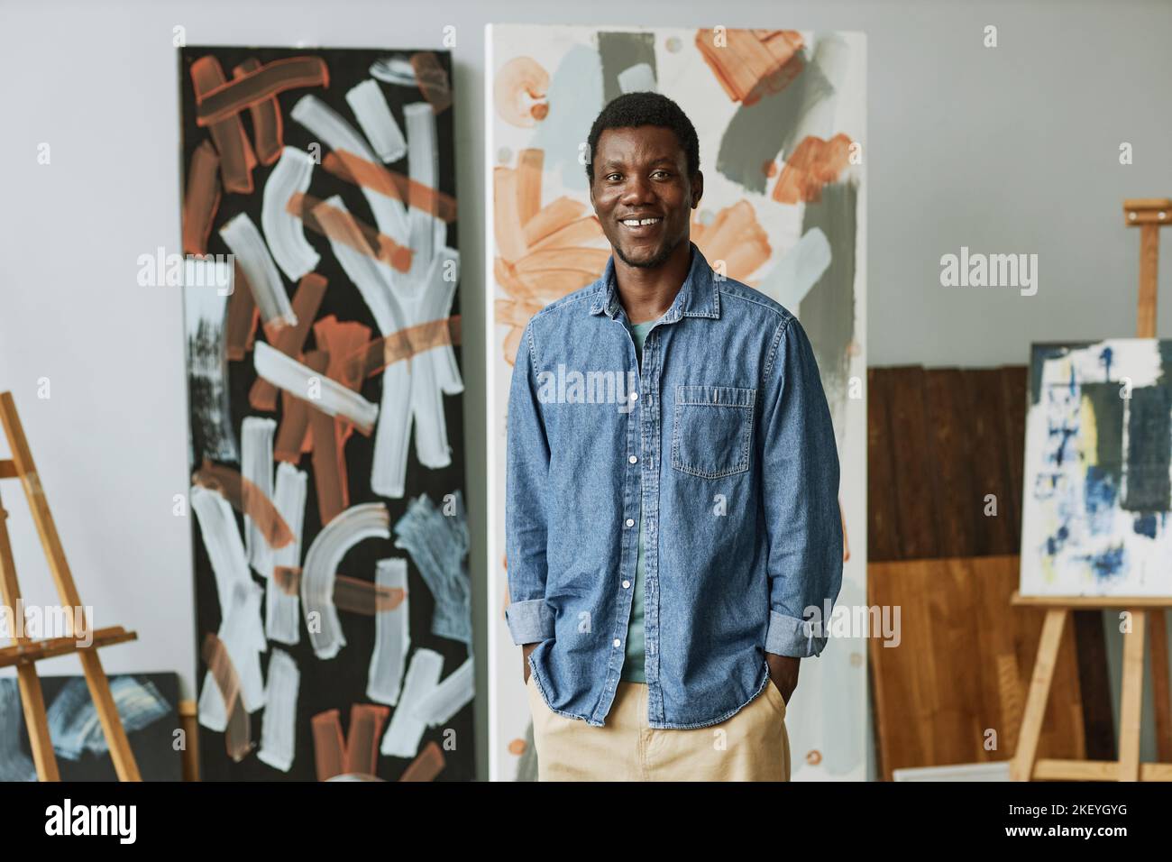 Junger lächelnder afroamerikanischer Handwerker in Casualwear, der auf dem Hintergrund abstrakter Gemälde im Studio oder Klassenzimmer steht Stockfoto