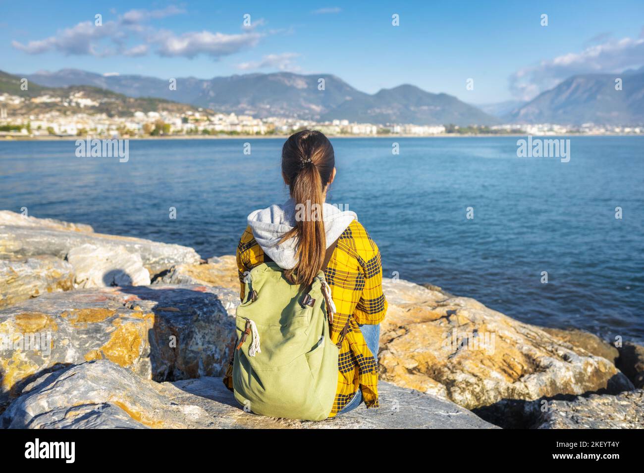Junge Reisende Frau mit Rucksack sitzt mit dem Rücken zur Kamera auf dem Stein am Meer und schaut in die Ferne Stockfoto