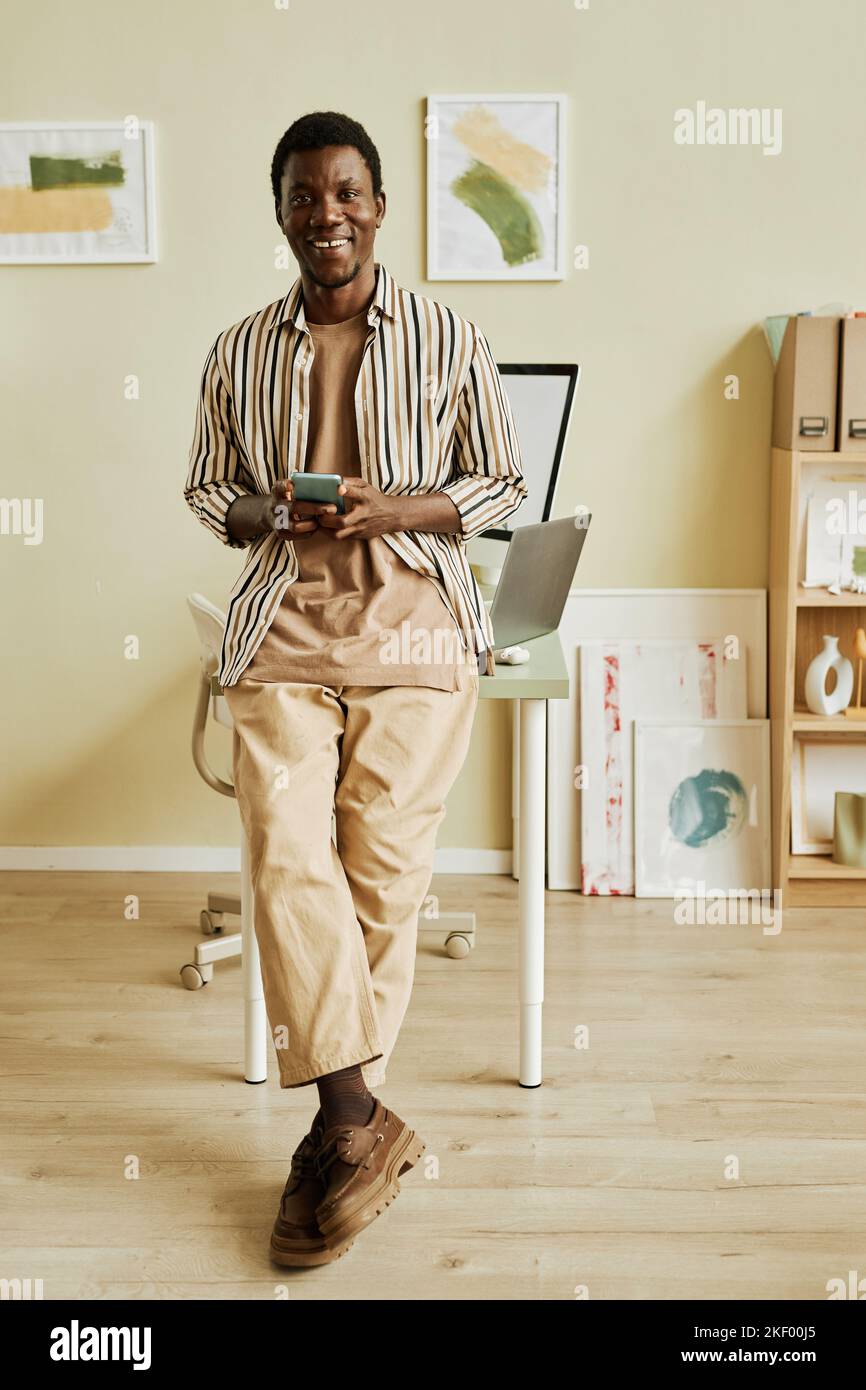 Fröhlicher junger kreativer Freiberufler in Casualwear mit Smartphone, während er mit Desktop-Computern im Büro gegen den Arbeitsplatz steht Stockfoto