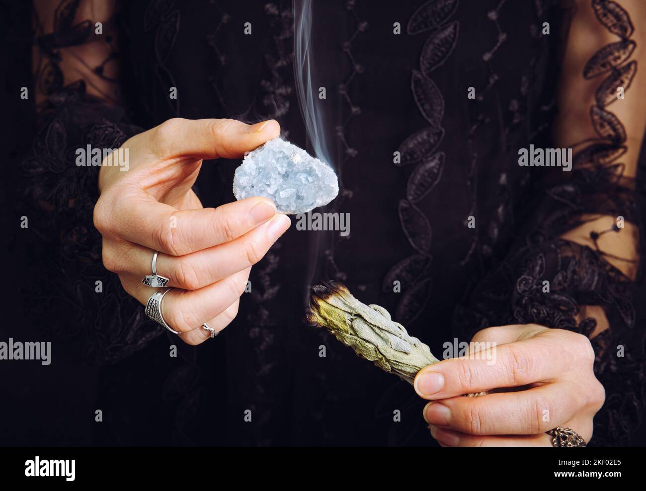 Nahaufnahme einer Frau in schwarzem Spitzenkleid, die den Edelstein eines blauen Himmelsteins aus Kristall reinigt, indem sie das weiße Salbei-Bündel verwischt. Entfernen Sie negative Energie. Stockfoto
