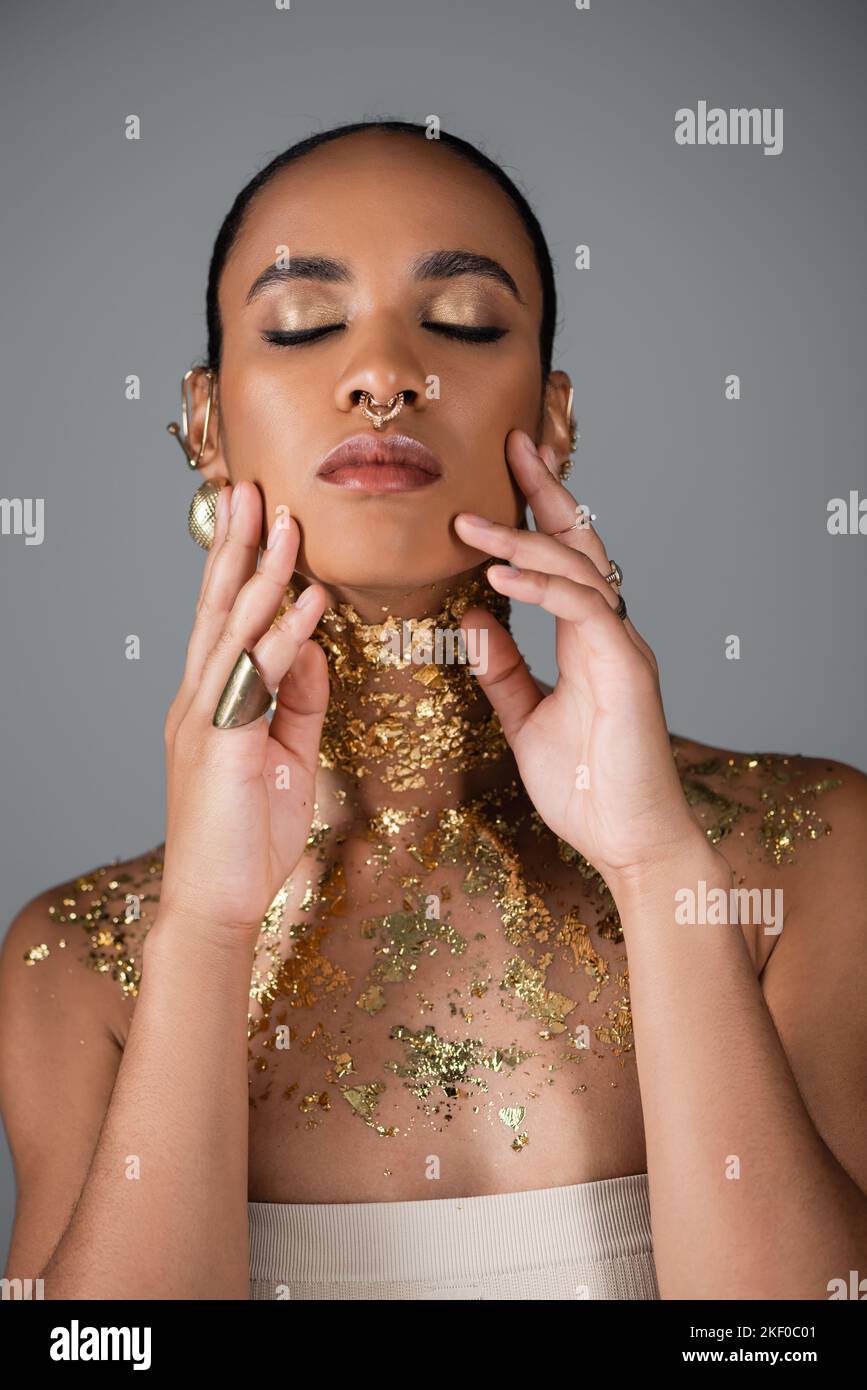Hübsche afroamerikanische Frau mit goldenem Make-up und Folie auf der Brust berühren Wangen isoliert auf grau, Stock Bild Stockfoto
