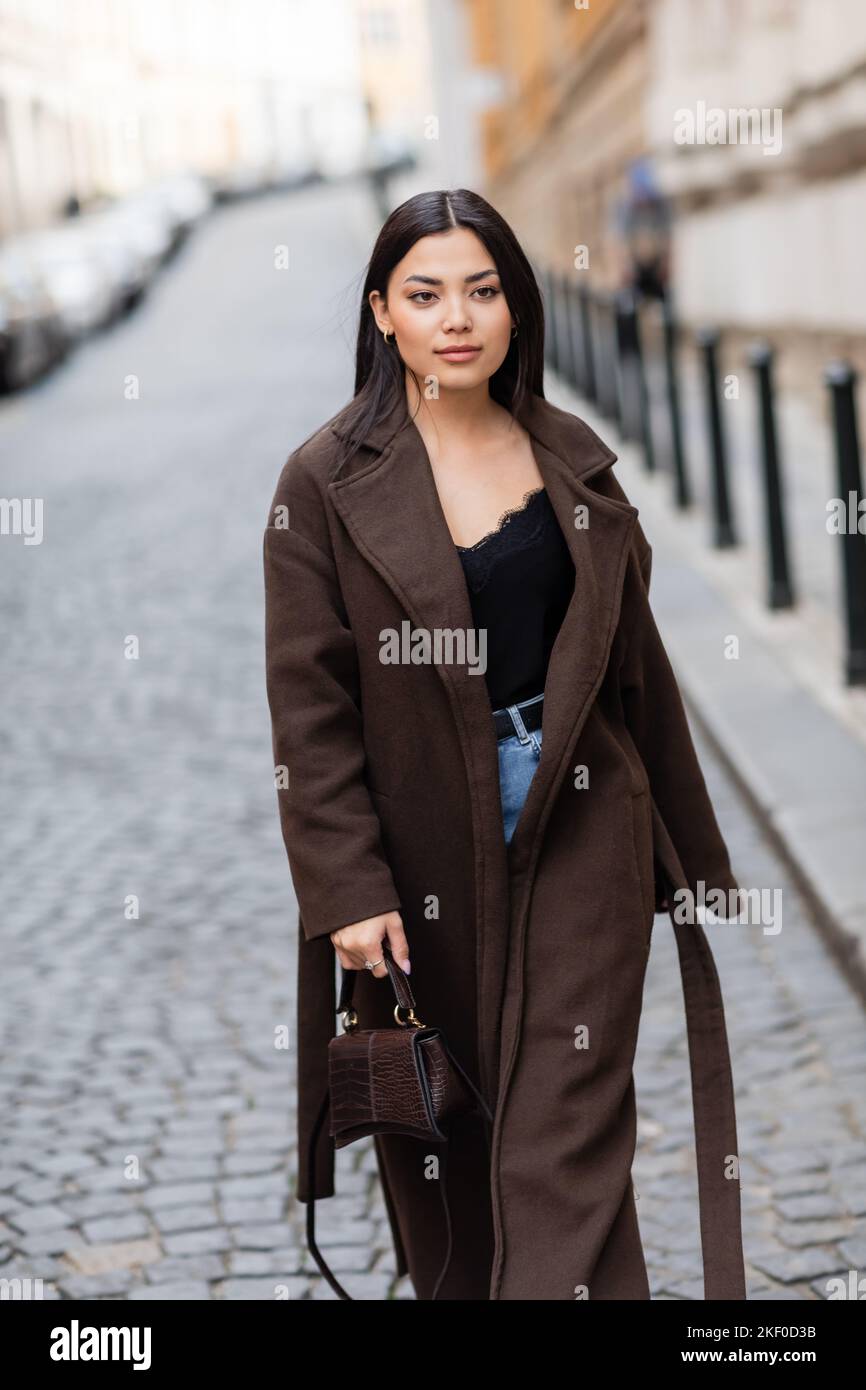 Stilvolle Brünette Frau in braunem Mantel halten Handtasche beim Spaziergang auf der Straße in prag, Stock Bild Stockfoto