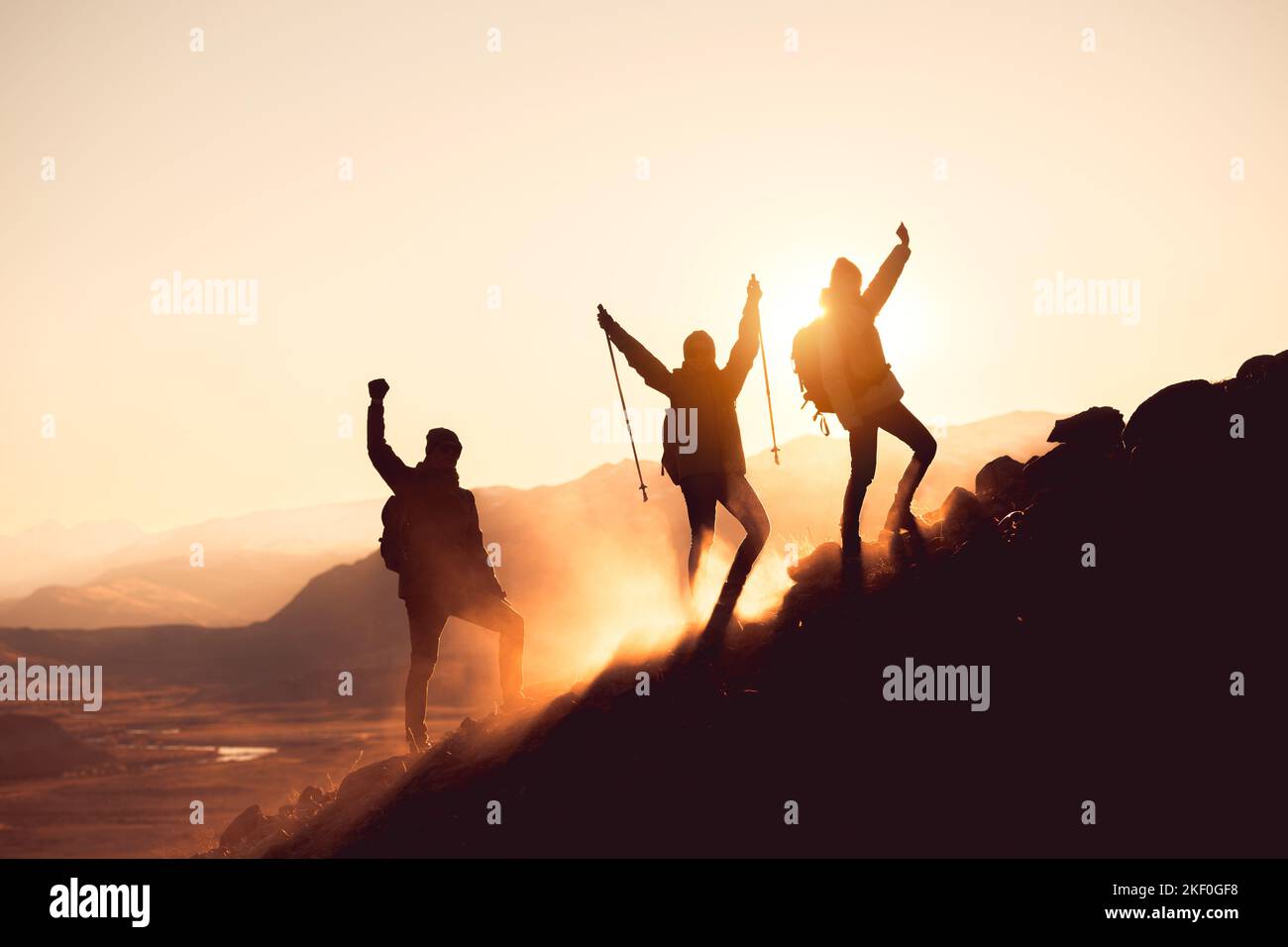 Silhouetten von drei glücklichen Wanderern, die mit erhobenen Armen am Berghang stehen Stockfoto