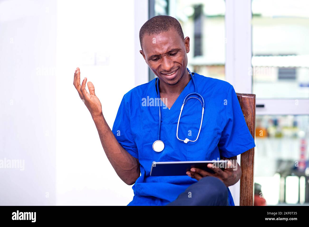 Junge ernsthafte männliche Arzt in blauer Uniform sitzt auf einem Stuhl arbeiten auf digitalen Tablet. Stockfoto