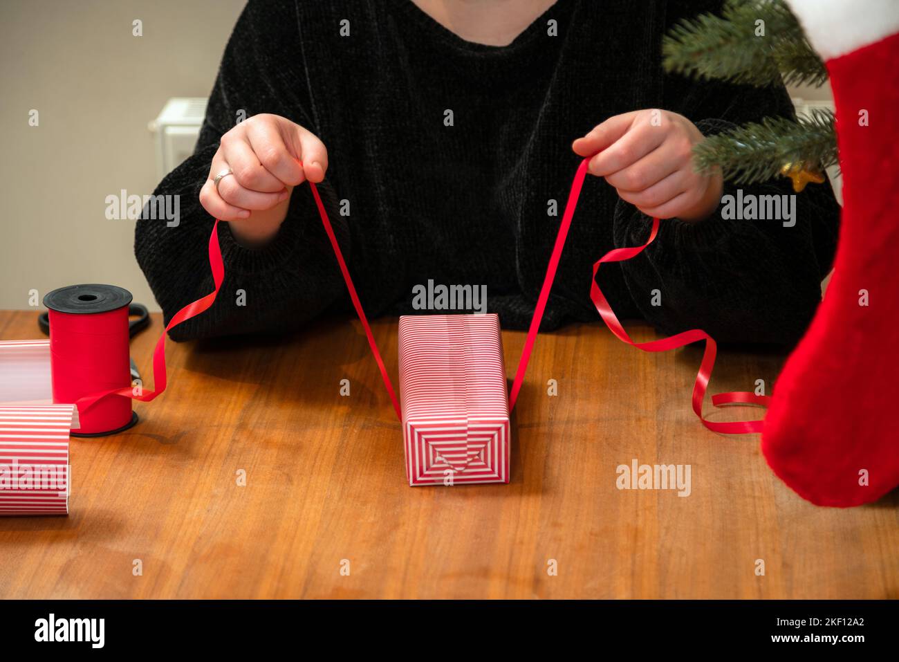 Frau verpackt ein Geschenk in abgestreiftes Papier mit rotem Band. Hände, die eine Schachtel in das rot-weiße weihnachtspapier packen. Stockfoto