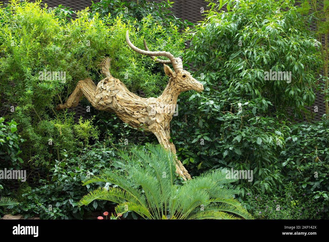 Ein Kunstwerk eines hölzernen Impalas, das von links vor einer buschigen Hecke herunterspringt. Stockfoto