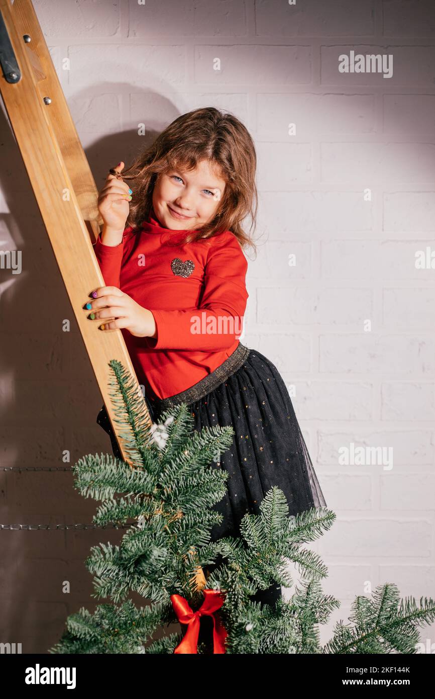 Kokett Mädchen posiert auf Holztreppen in festlicher Lage in der Nähe von weihnachtsbaum. Glückliches kleines Kinderporträt. Schöne Fichte für Innenarchitektur und Stockfoto
