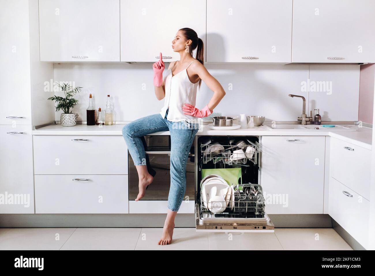 Ein Hausfrau-Mädchen in rosa Handschuhen sitzt nach der Reinigung des Hauses müde in der Küche.in der weißen Küche hat das Mädchen das Geschirr gewaschen und ruht sich aus Stockfoto