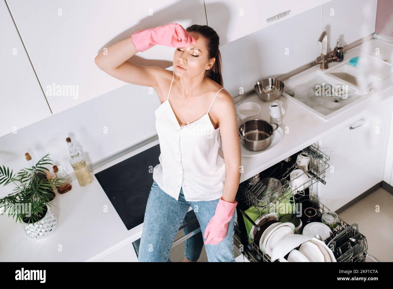 Ein Hausfrau-Mädchen in rosa Handschuhen sitzt nach der Reinigung des Hauses müde in der Küche.in der weißen Küche hat das Mädchen das Geschirr gewaschen und ruht sich aus Stockfoto