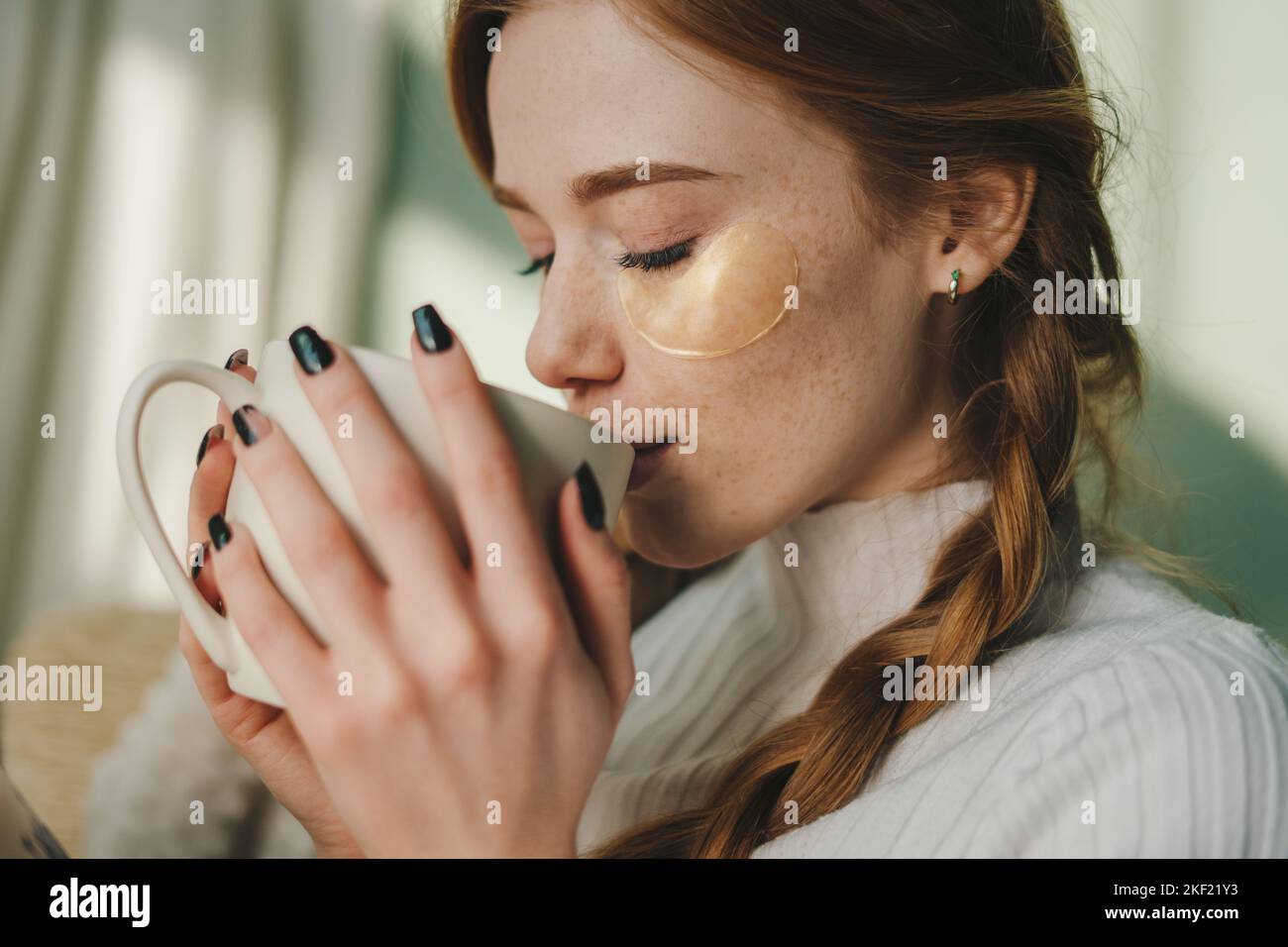 Porträt einer schönen Sommersprossen-Frau mit Augenflecken, die sich auf dem gemütlichen Sofa ausruhen und eine Tasse Tee trinken, um das Wochenende zu genießen. Unter Augenflecken. Freizeit Stockfoto