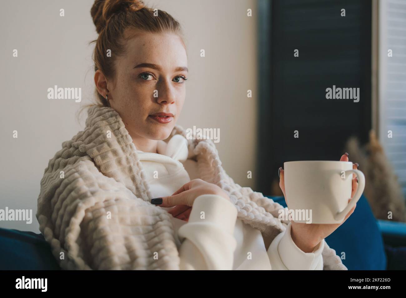 Porträt einer jungen kranken Frau, die auf die Kamera schaut, die auf dem Sofa sitzt und Tee trinkt. Tasse Tee, Kaffee und Chill. Freizeit genießen, Wochenende zu Hause Stockfoto