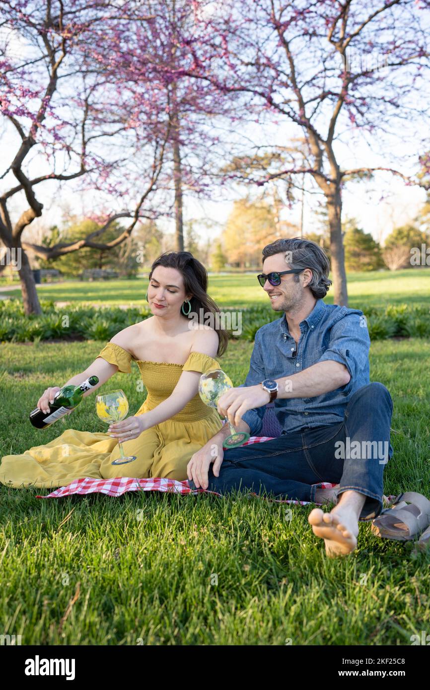 Ein Paar auf einer karierten Picknickdecke im Forest Park mit einer Flasche Wein und roten Knospenbäumen dahinter. Stockfoto