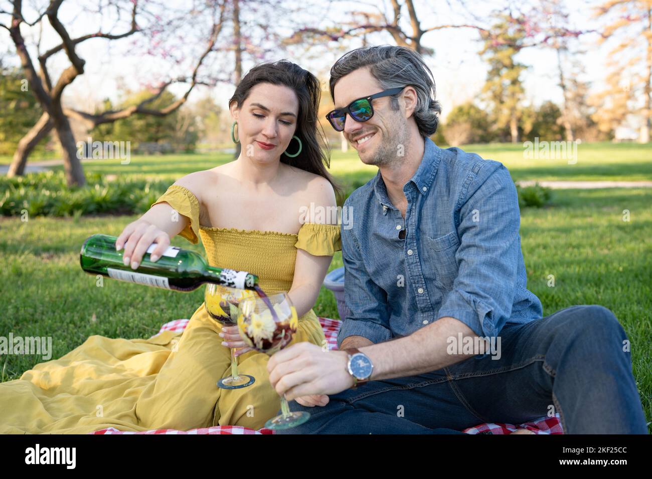 Ein Paar auf einer karierten Picknickdecke im Forest Park mit einer Flasche Wein und roten Knospenbäumen dahinter. Stockfoto