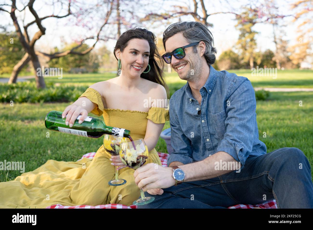 Ein Paar auf einer karierten Picknickdecke im Forest Park mit einer Flasche Wein und roten Knospenbäumen dahinter. Stockfoto