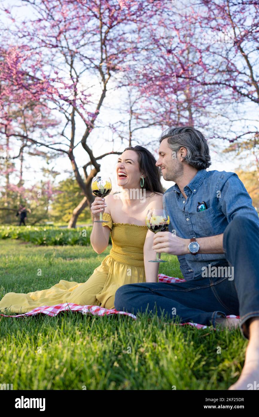 Ein Paar auf einer karierten Picknickdecke im Forest Park mit einer Flasche Wein und roten Knospenbäumen dahinter. Stockfoto