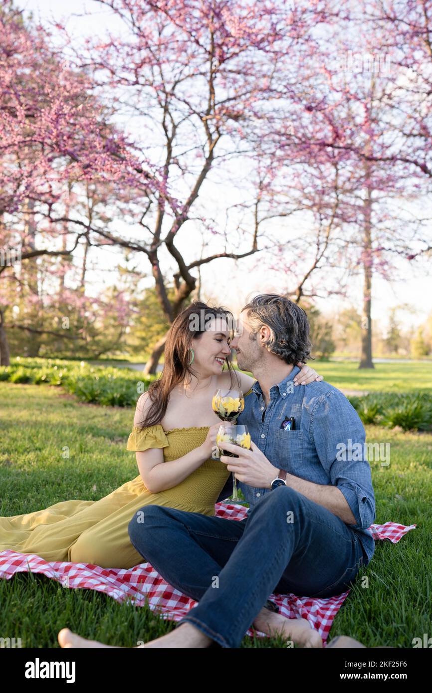 Ein Paar auf einer karierten Picknickdecke im Forest Park mit einer Flasche Wein und roten Knospenbäumen dahinter. Stockfoto