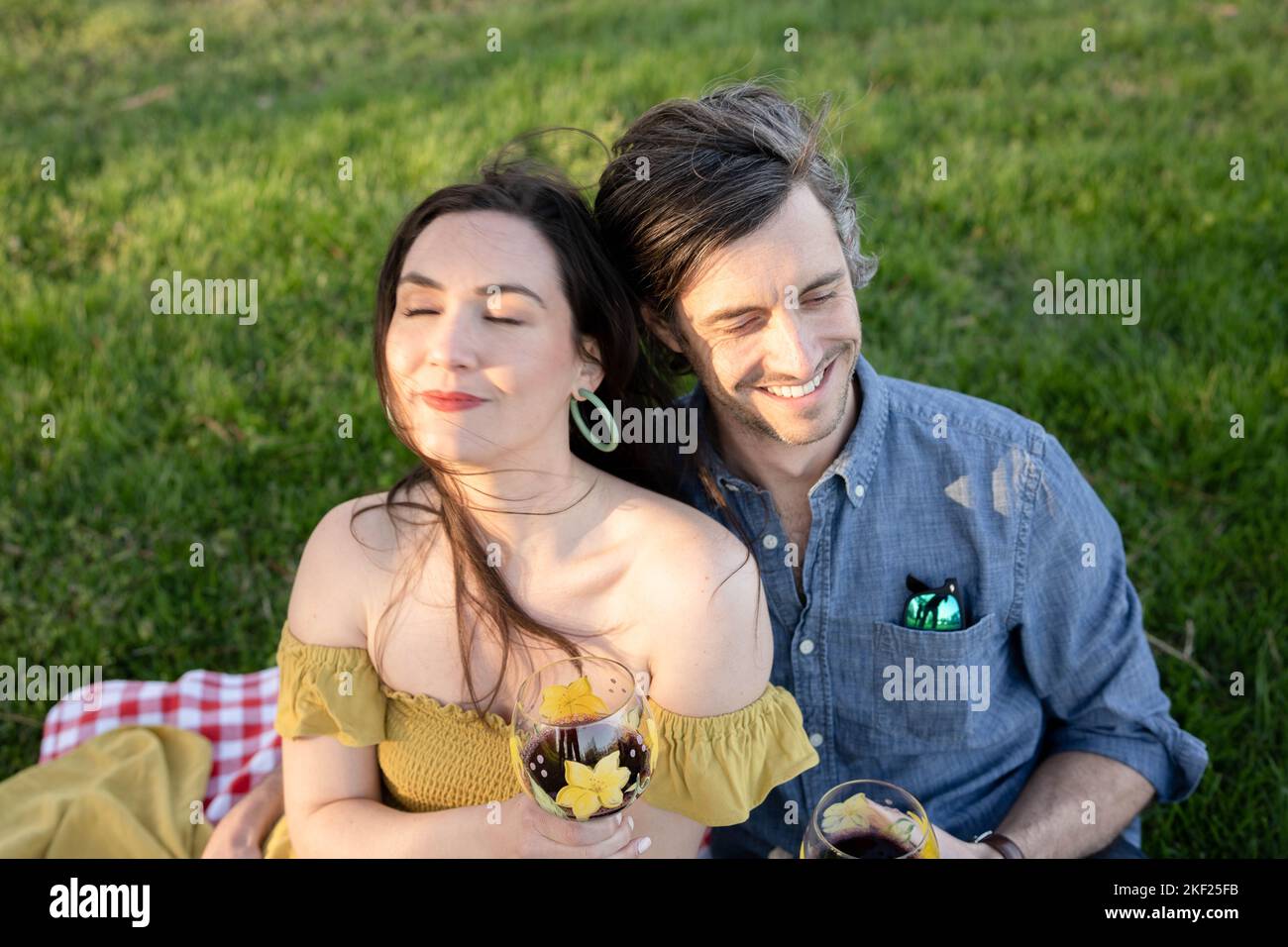 Ein Paar auf einer karierten Picknickdecke im Forest Park mit einer Flasche Wein und roten Knospenbäumen dahinter. Stockfoto