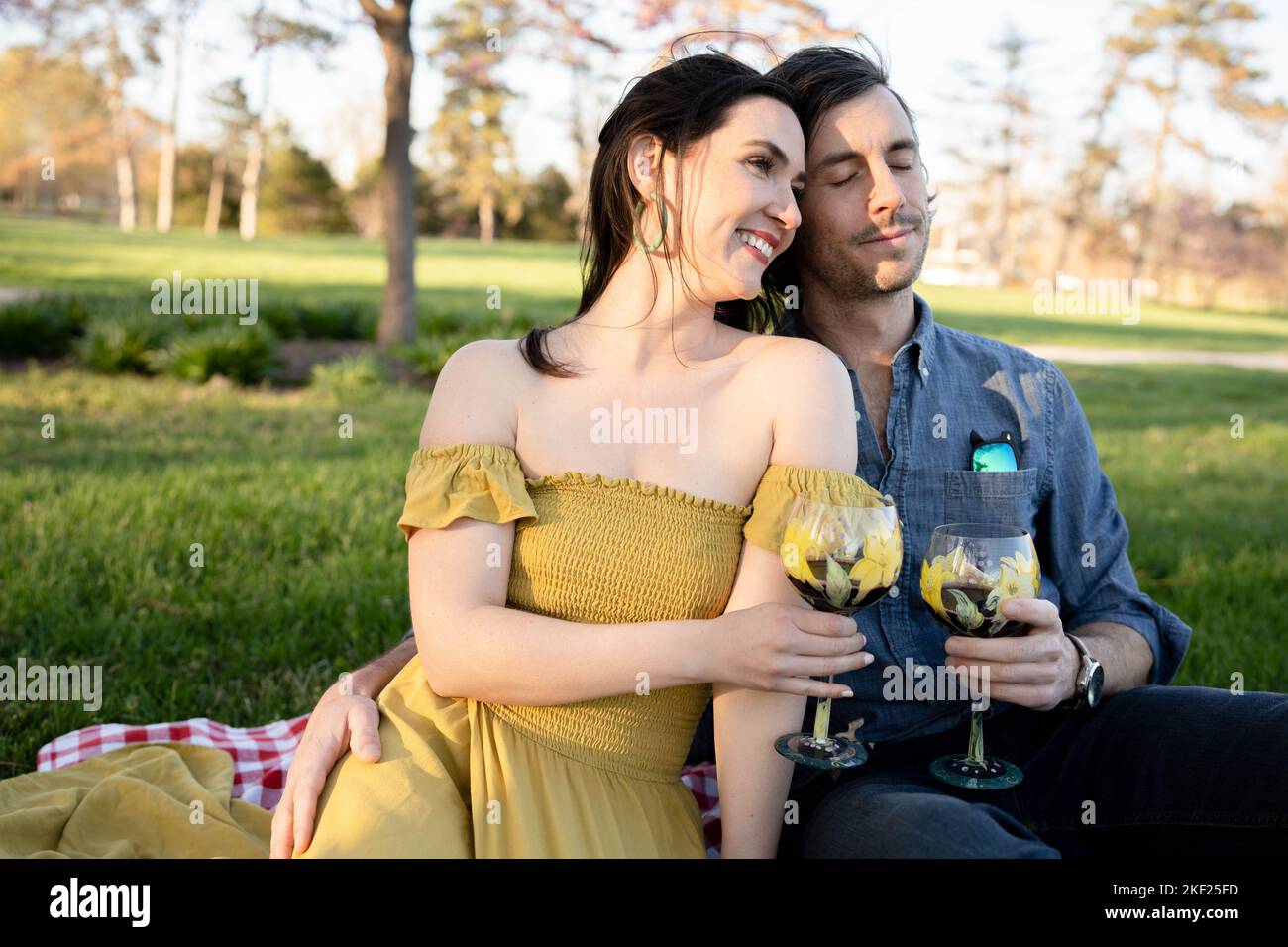 Ein Paar auf einer karierten Picknickdecke im Forest Park mit einer Flasche Wein und roten Knospenbäumen dahinter. Stockfoto
